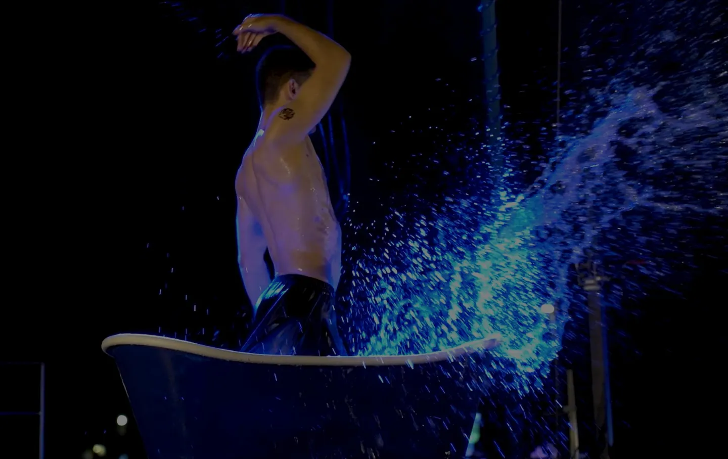 Acrobat on stage inside a bathtub, raising a wave of water lit in blue