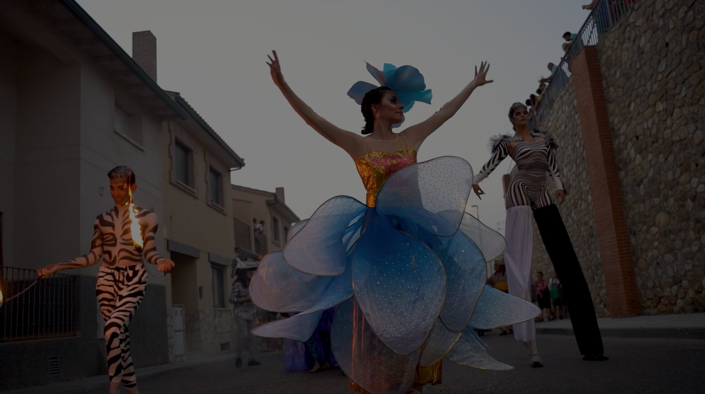 Street parade at dusk with a dancer dressed as a blue flower, a fire juggler and a stilt walker in a zebra costume