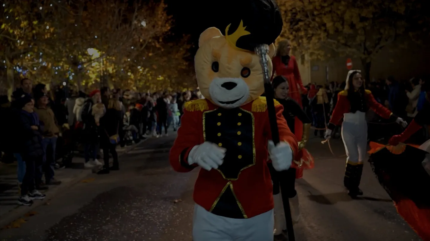 Desfile nocturno con un gran oso de peluche vestido de soldado, zancudos y público a ambos lados de la calle